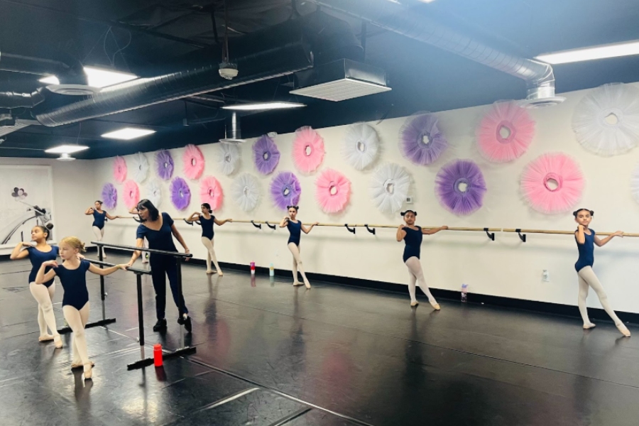 A ballet instructor guides young dancers in navy leotards as they practice at the barre in a studio with colorful tutu wall decorations.