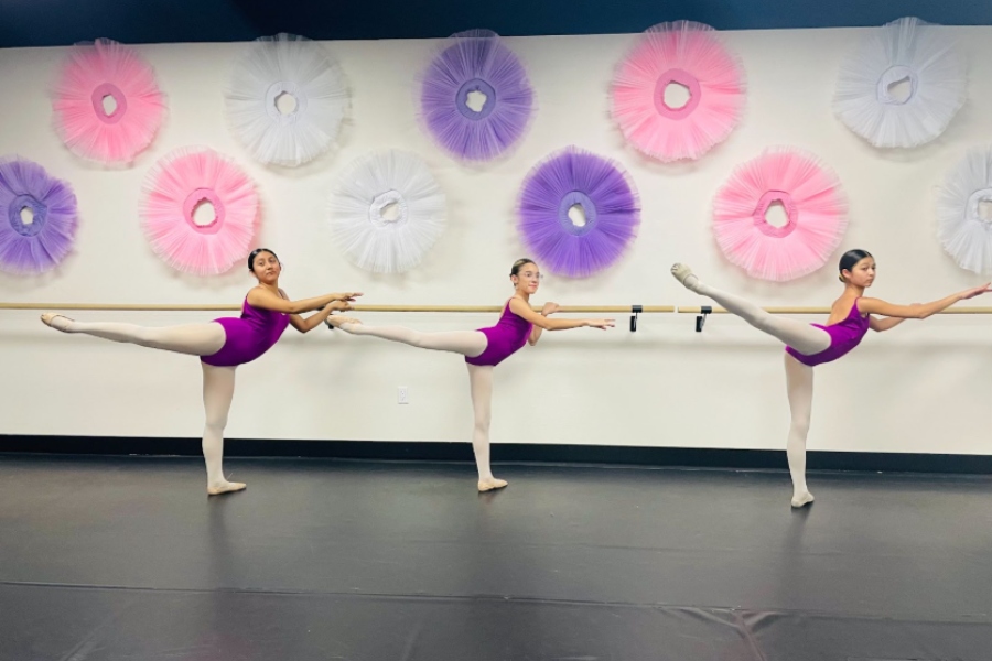Three young ballet dancers in purple leotards perform arabesques at a barre in a studio with colorful tutu wall decorations.