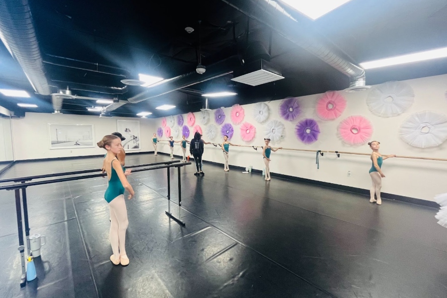 Young ballet students in teal leotards stand at the barre in a dance studio, guided by an instructor, with pastel tutu wall decorations in the background.