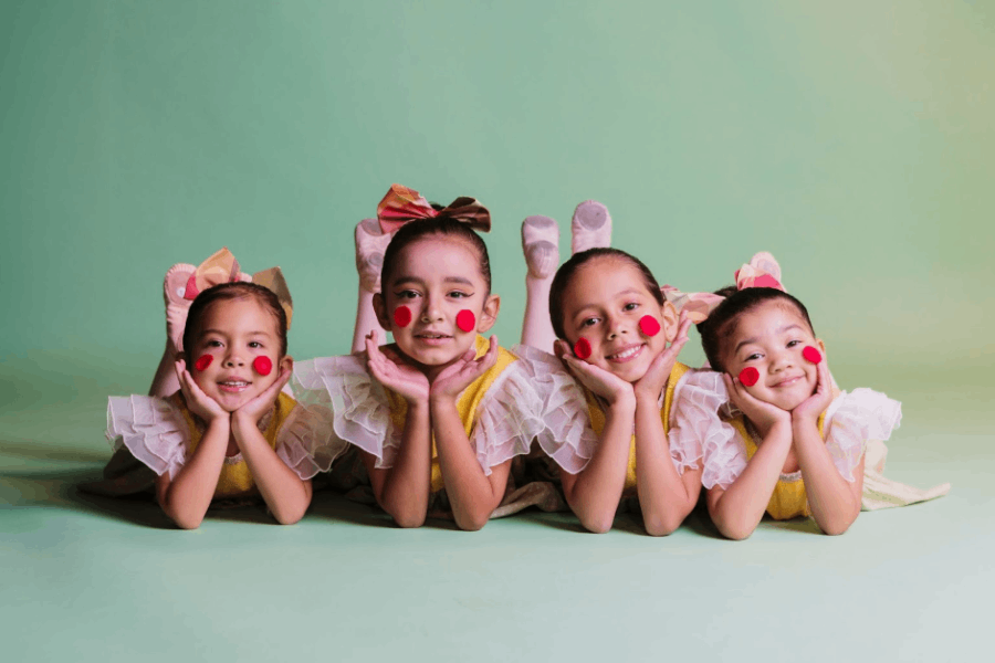 Four young girls dressed as clowns in colorful costumes and red cheek makeup lie on their stomachs smiling against a green backdrop.